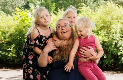 En kvinna med glasögon omgiven av tre blonda barn. De är utomhus med grönska i bakgrunden.