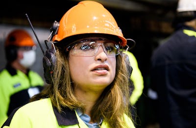 En kvinna med orange hjälm, skyddsglasögon och kläder med hög synlighet tittar framåt. Hon har ett radioknset och befinner sig i en industriell miljö med människor i bakgrunden.