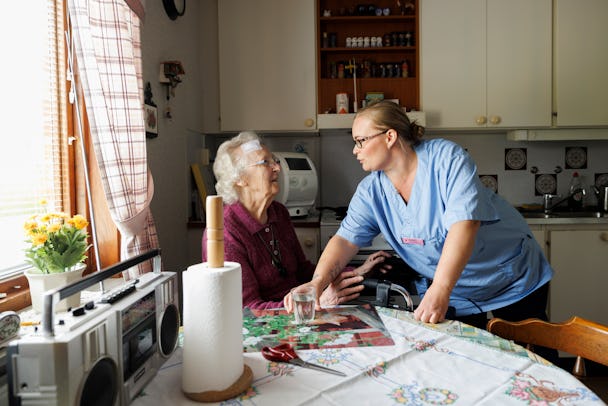 En vårdare i blå uniform hjälper en äldre kvinna vid ett köksbord. På bordet finns olika hushållsartiklar, som en radio och en pappersrulle. Köket har vita skåp och ett fönster med gardiner.