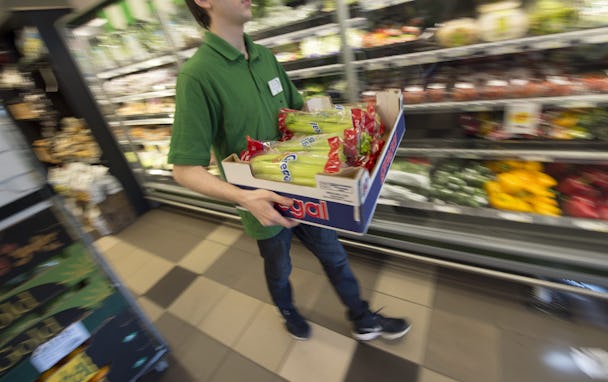 En person iklädd en grön skjorta bär en låda med förpackade snacks genom en gång i mataffären som innehåller grönsaker och annan färskvaruproduktion.