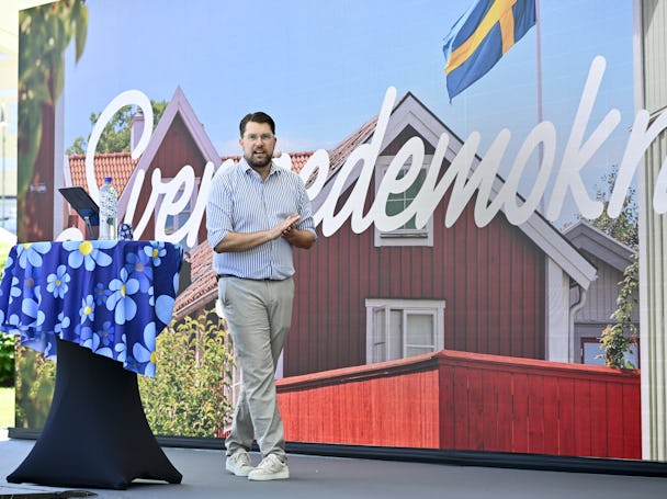 En man står på scenen och applåderar. I bakgrunden finns svensk text och en husbild. En hylla med en blomsterprydd duk och en vattenflaska står bredvid honom.