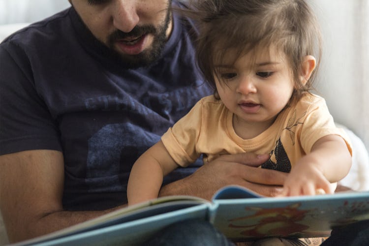En man håller ett litet barn på sitt knä. Barnet håller i och tittar på en bilderbok medan mannen ser på.