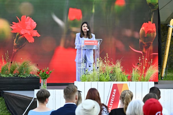 Nooshi Dadgostar talar vid ett podium med en bakgrund av röda nejlikor. En publik lyssnar framför scenen som är dekorerad med växter och blommor.