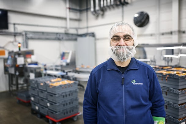 En arbetare i en livsmedelsproduktionsanläggning som bär hårnät och skäggskydd, med förpackningsmaskineri i bakgrunden.