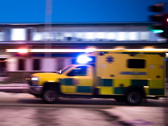 En gul och grön ambulans med blinkande ljus kör snabbt på en stadsgata i skymningen.