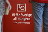 Röd skylt med vit text som lyder "Vi får Sverige att fungera, LOs 30e kongress." Två personer står bredvid skylten, en håller i en kaffekopp.
