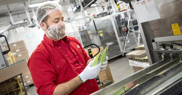 En arbetare i röd uniform och hårnät inspekterar förpackningar i en fabrik, stående bredvid maskiner och transportband.