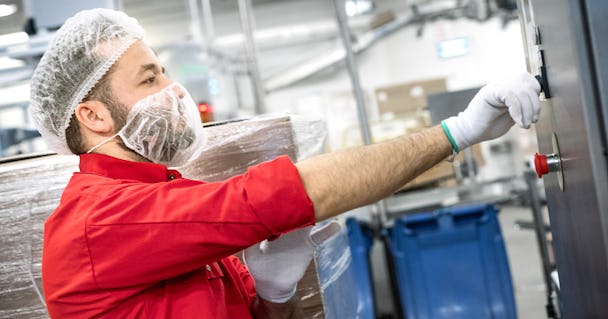 En person i röd skjorta, hårnät och handskar använder maskiner i en industriell anläggning.