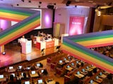 Foto inifrån en fullsatt kongressal, med prideflaggans färger lagda ovanpå. Bilden är ett montage.