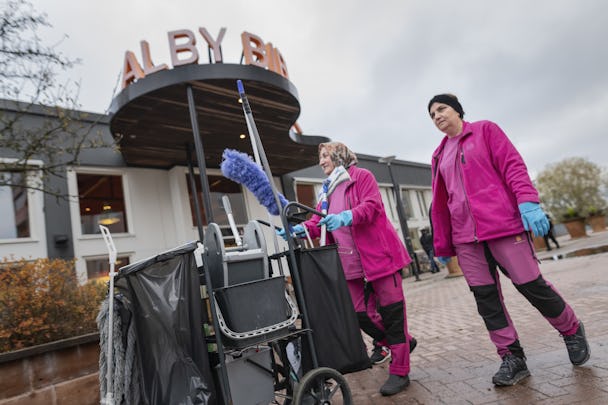 Zeliha Bulduk och Gülüzar Atik går utomhus i rosa arbetskläder med en städvagn under en stor skylt där det står ”Alby”.