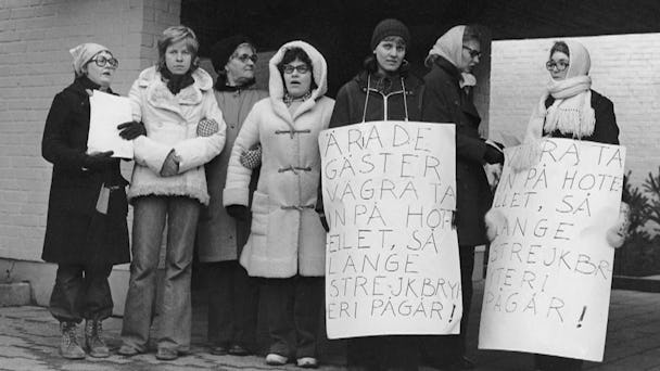 Svartvit arkivbild från 1974 på strejkande städerskor med plakat. På ett av dem står det ”Ärade gäster vägra ta in på hotellet så länge strejkbryteri pågår!”