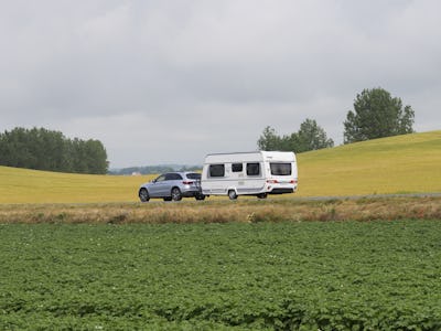 Ett fordon som drar en husvagn som rör sig på en väg med omgivande fält och en mulen himmel.
