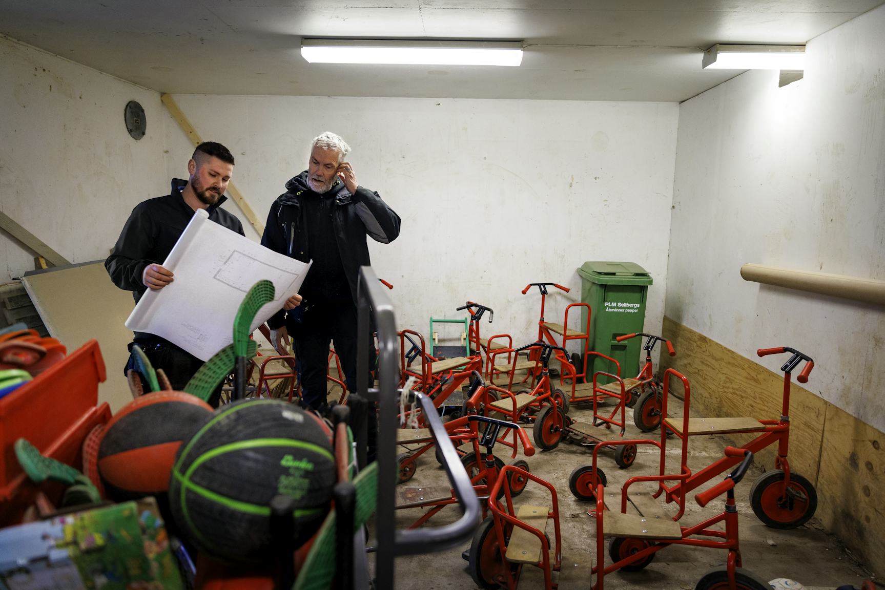 Johan Söderberg och Rickard Jönsson studerar en ritning i ett stökigt skyddsrum med utspridda trehjulingar.