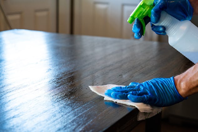 En person i blå handskar använder en sprejflaska och trasa för att rengöra ett träbord.