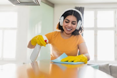 En kvinna med hörlurar och gula handskar rengör en yta med en sprayflaska och trasa.