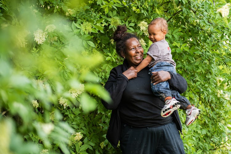 Fahima Doodi i svarta kläder håller sitt barn framför grönt lövverk; båda ler.