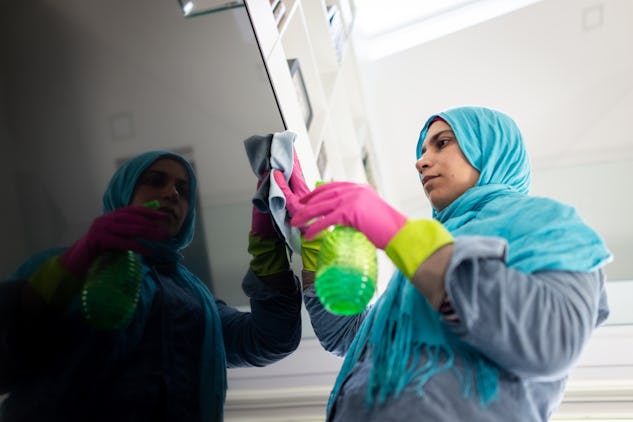 En person med en blå huvudduk och rosa handskar rengör en spegelyta med en trasa och en grön sprayflaska.