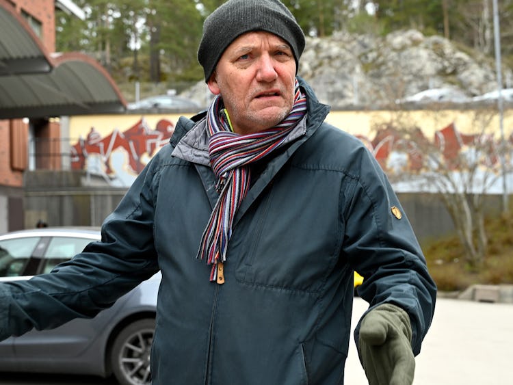 En man i vinterrock, halsduk och handskar står utomhus.