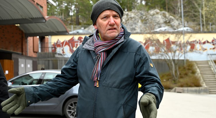 En man i vinterrock, halsduk och handskar står utomhus.