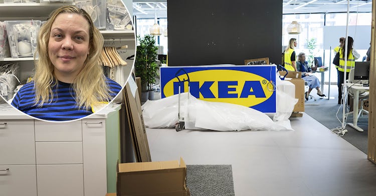 En kvinna med blont hår står i ett förvaringsområde. Bakom henne syns en IKEA-skylt tydligt i en kontorsmiljö där flera personer inte läser varselvästar deltar i en diskussion.