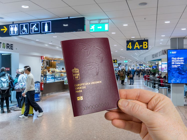 Monterad bild. En hand håller i ett svensk pass av 2022 års modell. I bakgrunden en massa folk någonstans på Arlanda flygplats.