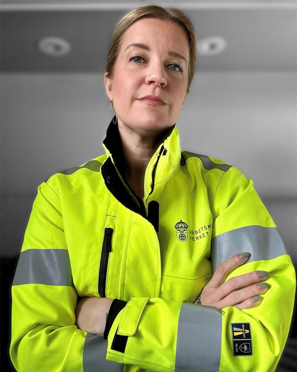Jenny Bengtsson tittar in i kameran med armarna i kors, bärandes en gul varseljacka med Arbetsmiljöverkets logotyp.