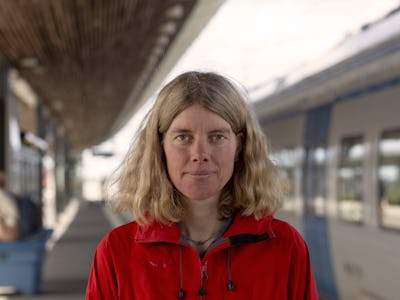 Vänsterpartiets Anna Sehlin i röd jacka på en pendeltågsstation framför ett vitt pendeltåg med blå dörrar.