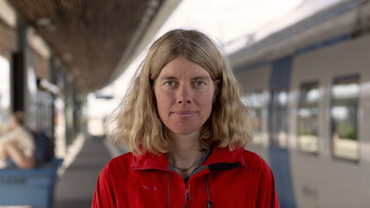 Vänsterpartiets Anna Sehlin i röd jacka på en pendeltågsstation framför ett vitt pendeltåg med blå dörrar.