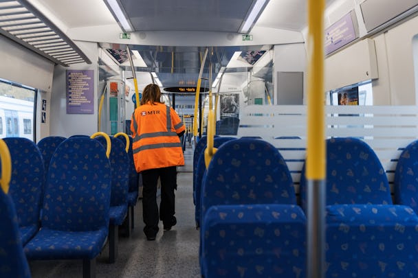 Ryggen på en anställd som går i ett pendeltåg. Personen bär en orange varselväst med texten MTR.