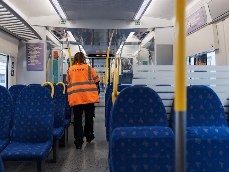 Ryggen på en anställd som går i ett pendeltåg. Personen bär en orange varselväst med texten MTR.