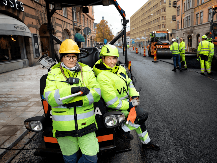 Signalvakterna Annica Björk och Jeanette Lundberg i full mundering av gula varselkläder vid en gata där asfalten läggs om. De står lutade mot en asfaltsmaskin.