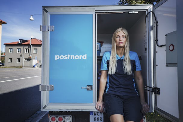 Brevbäraren Emilia Leijon Amrén vid en Postnord-bil, iklädd Postnords blåa uniform en sommardag.