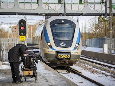 Ett pendeltåg kommer inkörandes till Kallhäll station. I förgrunden, på perrongen, står en person med en barnvagn.