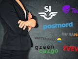 En anonym affärskvinna med korsade armar som står framför en vägg med olika svenska företagslogotyper inklistrade.