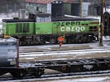 En arbetare i synliga kläder går förbi tågvagnar på snötäckta spår med ett grönt lastlokomotiv i bakgrunden.