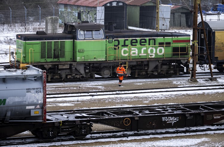 En arbetare i synliga kläder går förbi tågvagnar på snötäckta spår med ett grönt lastlokomotiv i bakgrunden.