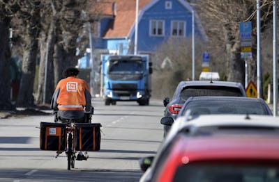 En brevbärare på cykel från Citymail cyklar längs en gata och är på väg att möta en blå lastbil från Postnord.