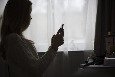 En ung kvinna sitter vid ett fönster och håller en smartphone. Hennes silhuett är delvis belyst av ljuset som kommer genom de tunna gardinerna.