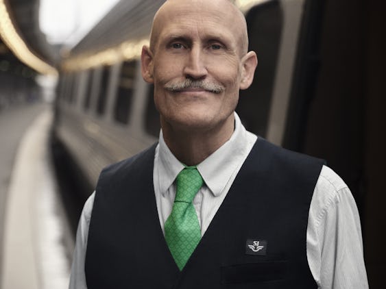 Lars Fuhre iklädd en vit skjorta, grön slips och svart väst med "SJ"-logga, står framför ett tåg på en perrong.