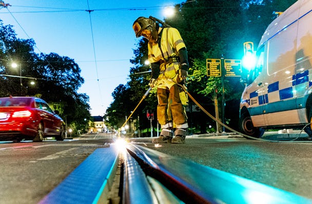 En arbetare iförd skyddsutrustning svetsar en spårvagnsskena på natten på en stadsgata. Trafik passerar förbi och en tjänstebil är parkerad i närheten.