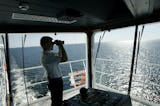 En person i uniform använder en kikare för att titta ut över havet från ett fartygs brygga.