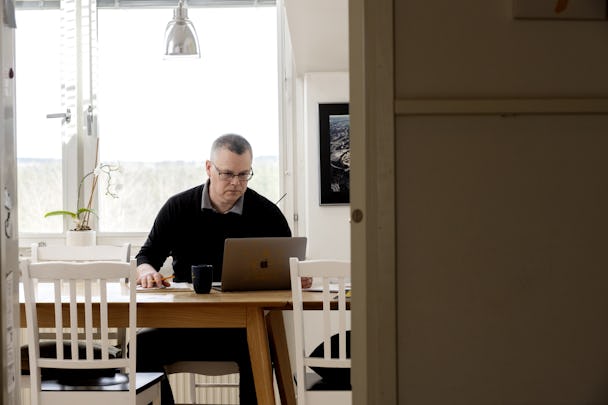 Patrik Nylund sitter vid sin bärbara dator på ett träbord. En mugg och en krukväxt står på bordet. Över bordet hänger en ljusarmatur, och i bakgrunden finns ett stort fönster.