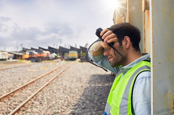 Man i neongul väst lutar sig mot metallstruktur, skuggar sina ögon och håller en walkie-talkie. I bakgrunden syns järnvägsspår och flera tåg.