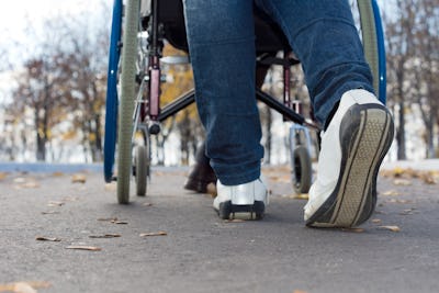 Närbild av en person i rullstol och en annan person som går bredvid på en utomhusväg med fallna löv.