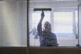 Person rengör ett fönster med en fönsterskrapa från insidan av ett rum, sett genom persienner.