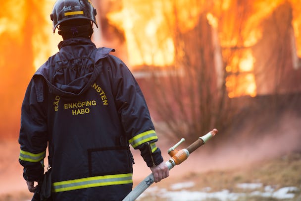 En brandman står med en slang framför ett brinnande hus.