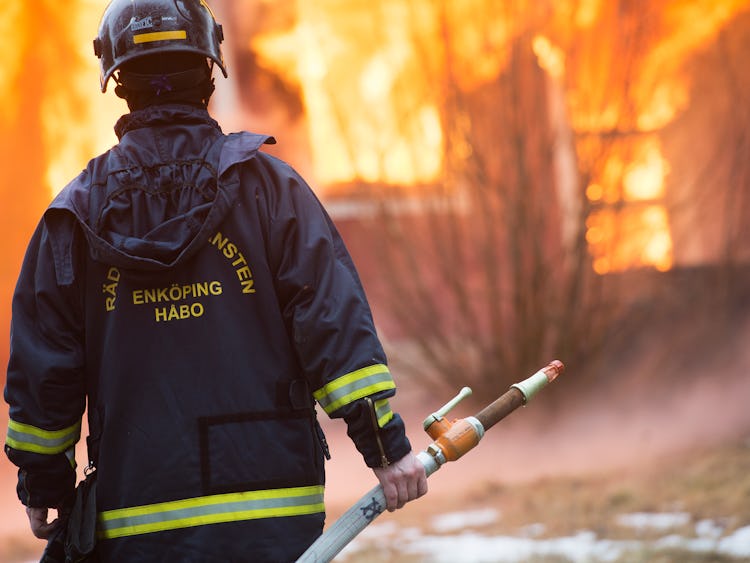 En brandman står med en slang framför ett brinnande hus.