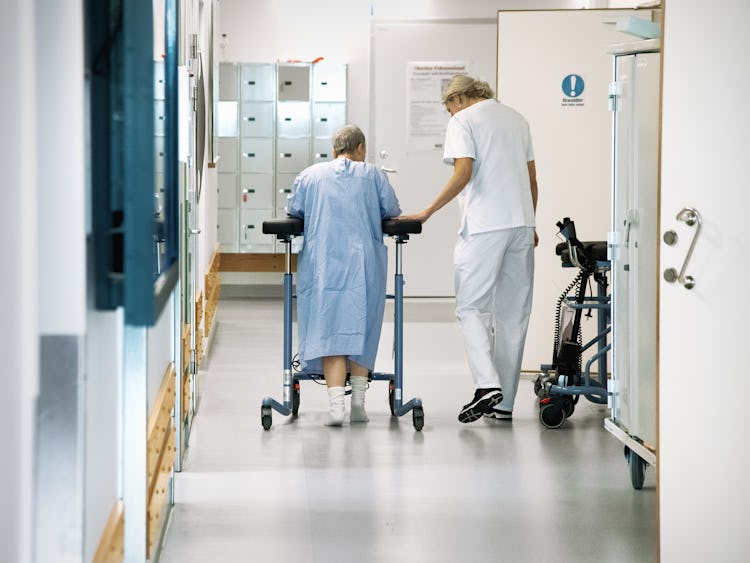 En kvinnlig patient med en rollator får hjälp av personal att ta sig fram i en korridor på ett sjukhus