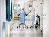 En kvinnlig patient med en rollator får hjälp av personal att ta sig fram i en korridor på ett sjukhus .