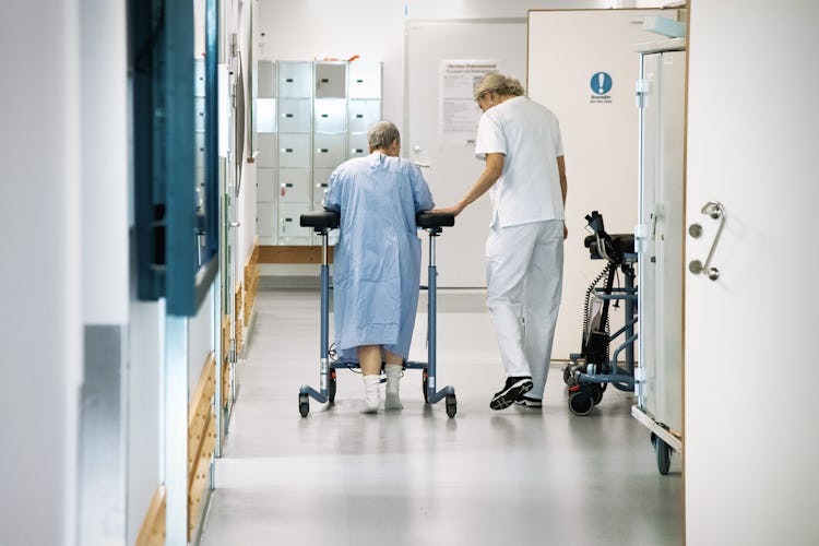 En kvinnlig patient med en rollator får hjälp av personal att ta sig fram i en korridor på ett sjukhus .